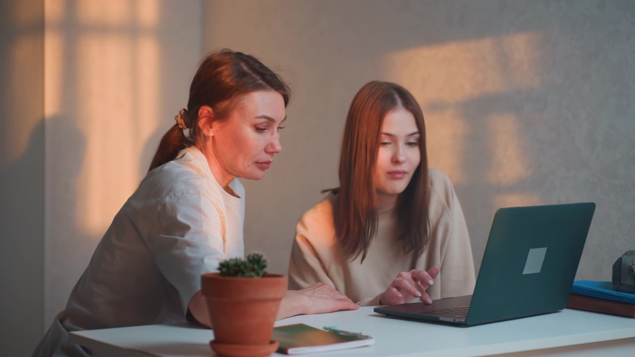 Sessione di tutoraggio a domicilio al laptop, insegnante/coach guida studente adolescente attraverso esercizi di grammatica in un accogliente ufficio domestico, calda luce solare serale cade sulla scrivania con pianta in vaso e quaderno aperto, concentrato.