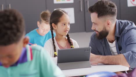 Diverse-male-teacher-and-happy-schoolchildren-using-tablet-in-school-classroom