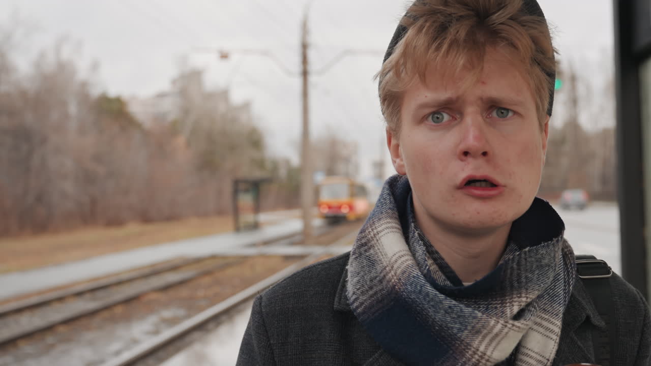 Junger weißer Kreativer steht an der Straßenbahn, Herbstregen, Schal und Mütze, Regenschirm in der Hand, nachdenklich blickend, Shotlist planend, bedecktes Licht, nasse Gleise und ferner Verkehr, stilvoller Mantel