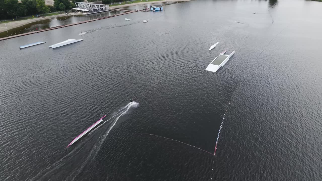High-resolution aerial drone footage of a kitesurf cutting across a calm lake near a watersport park, with floating ramps and obstacles visible in the water