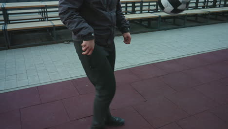 partial view of athlete in hoodie juggling ball with skillful precision during training in urban sport arena, with background featuring benches and cityscape