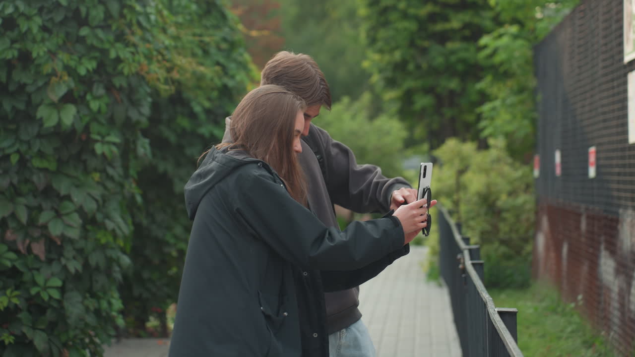 Giovane coppia che si scatta un selfie vicino a una recinzione, smartphone tenuto vicino mentre si appoggiano l'uno all'altro, espressioni gioiose e sguardo condiviso sullo schermo, sentiero del giardino esterno con segnaletica, giacche casual e movimento spontaneo