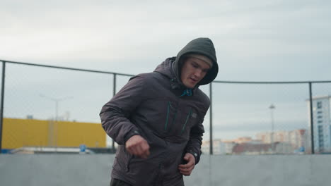 focused man in hoodie skillfully controlling soccer ball with precise footwork on sport arena, urban background features a yellow barrier, fence, and distant residential buildings