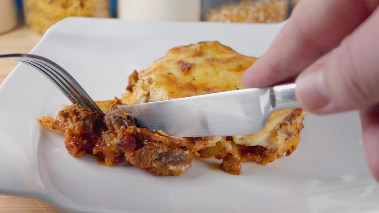 Primo piano al rallentatore di una porzione di lasagne appena sfornate su un piatto bianco con coltello e forchetta