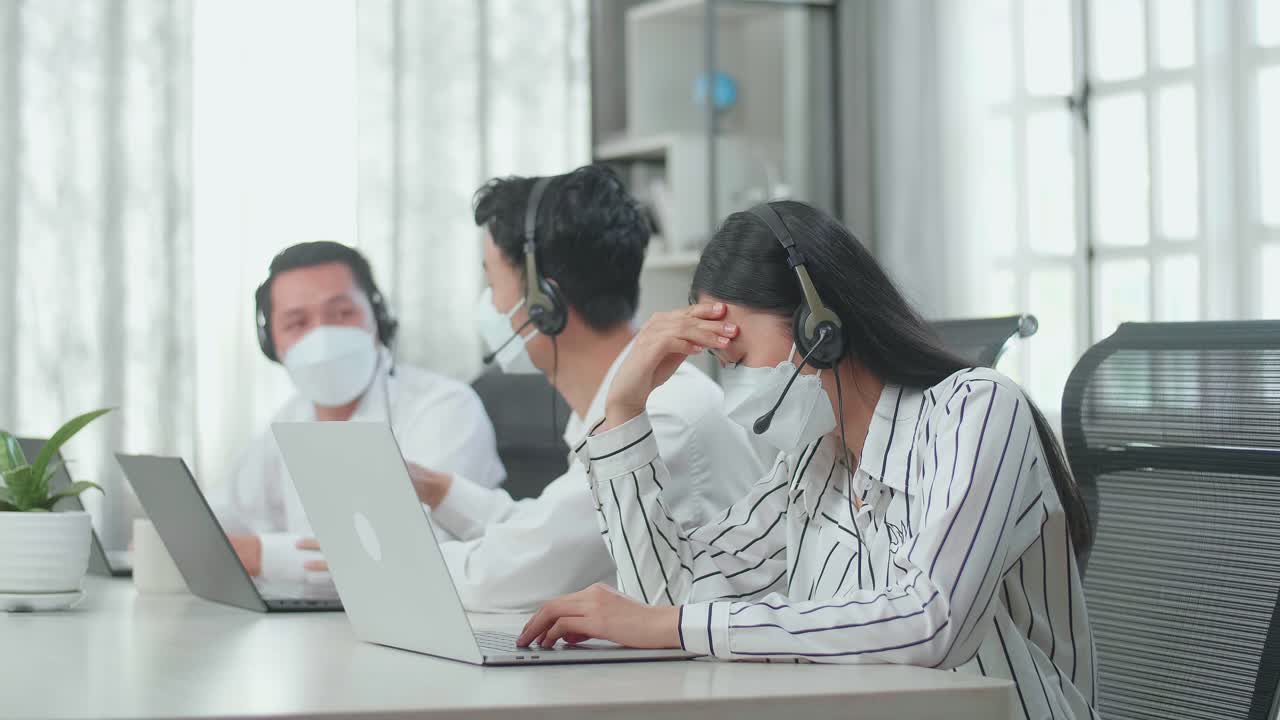 A Woman Of Three Asian Call Center Agents Wearing Headset And Mask Headache Because Two Of Her Colleagues Are Talking During Working At The Office