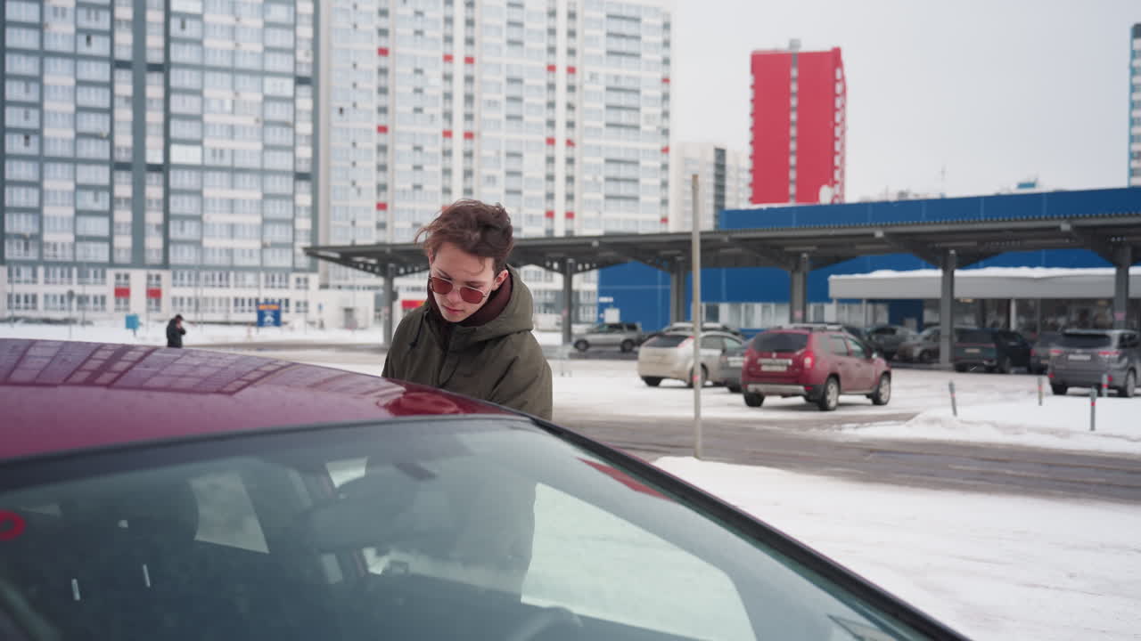 Taxi driver wearing winter jacket walking toward car looking at person in distance before entering vehicle with snow covering parking lot and tall office buildings visible in urban winter background