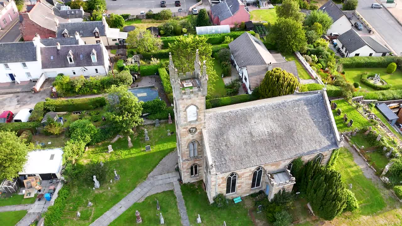 Drone footage circles a stone church with clock tower and lush garden in Rosemarkie, Highland, Scotland, under bright natural daylight