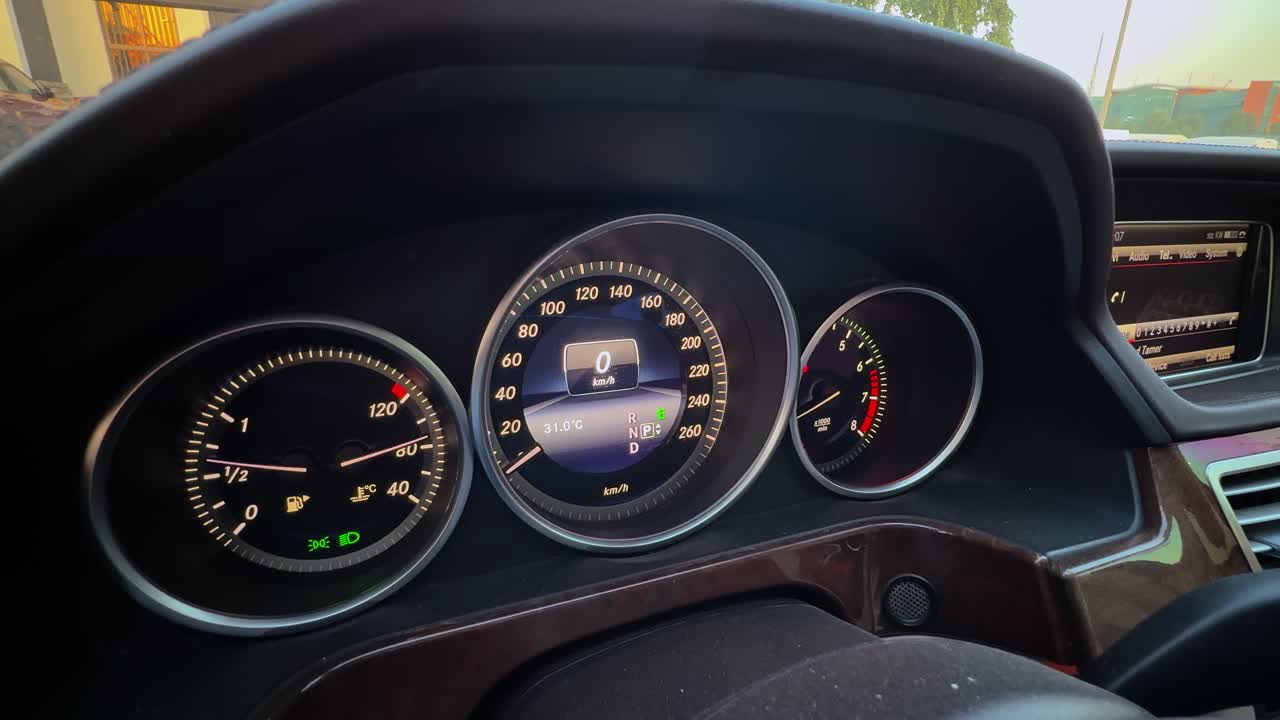 a high-end car instrument cluster. Digital speedometer shows zero km h during the day with illuminated gauges and polished wood trim accents in a modern vehicle cabin