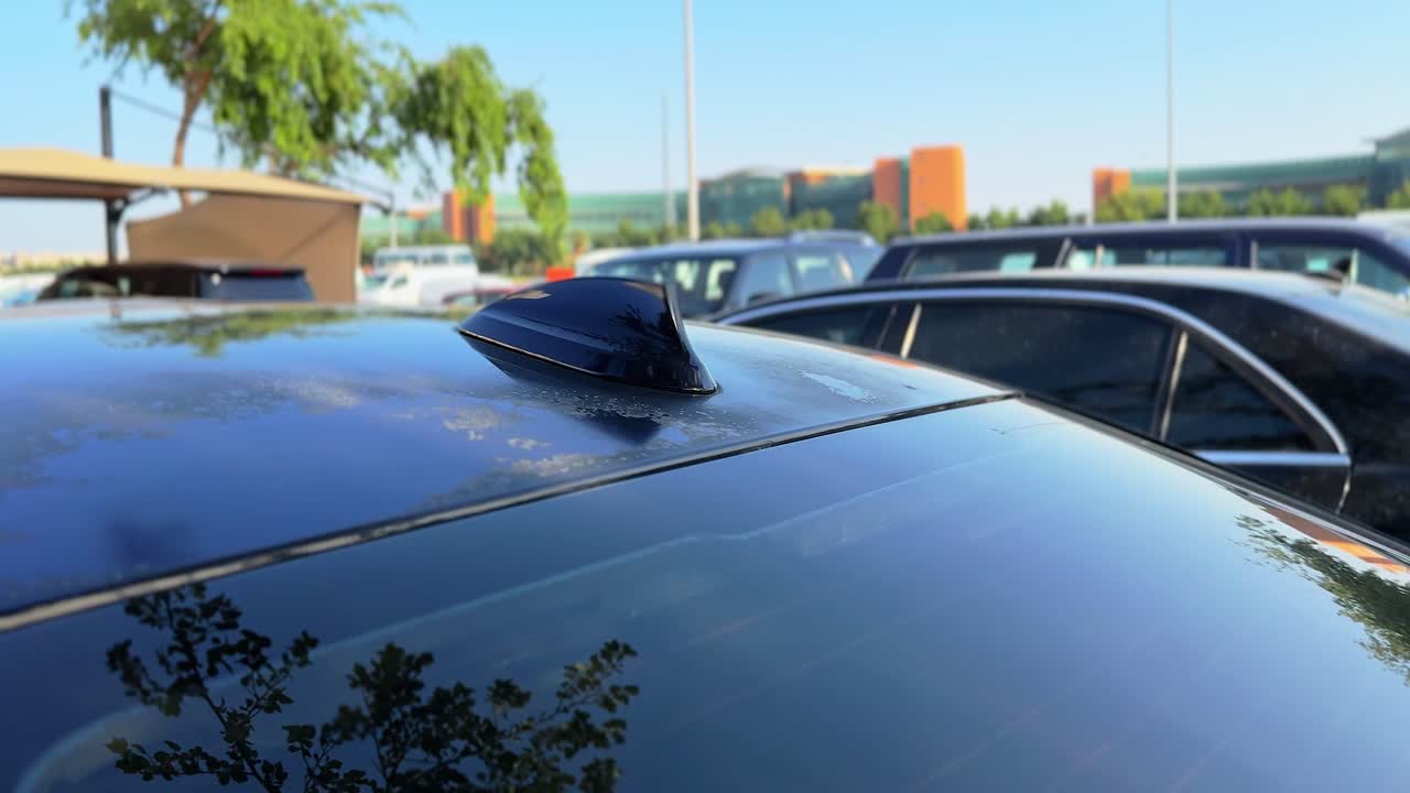 Un'antenna a pinna di squalo nera sul tetto di un'auto blu. Il veicolo è parcheggiato in un'area di sosta esterna soleggiata con edifici moderni e vegetazione sullo sfondo, in pieno giorno.