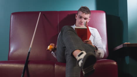 focused young man in white shirt and plaid pants sits on red sofa in billiard room, legs crossed, reading book while rolling orange billiard ball in hand. cue stick leans beside him