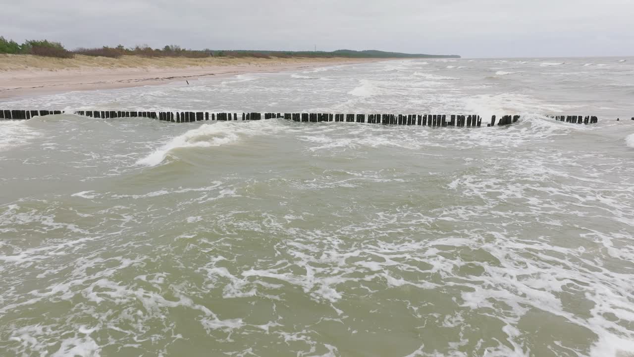 흐린 날 발트해 연안의 공중 전망, 오래된 목재 부두, 하얀 모래 해변, 해안에 부딪히는 큰 폭풍우, 기후 변화, 중간 드론 샷이 앞으로 낮게 움직입니다.