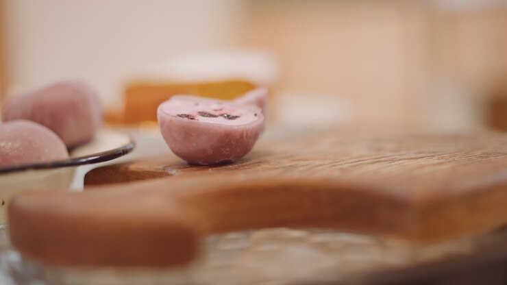 Pink mochi resting on wooden board in soft focus spoon beside slice, blurred pumpkin pie in background, pastel tones, studio tabletop composition, calm cozy mood for dessert showcase