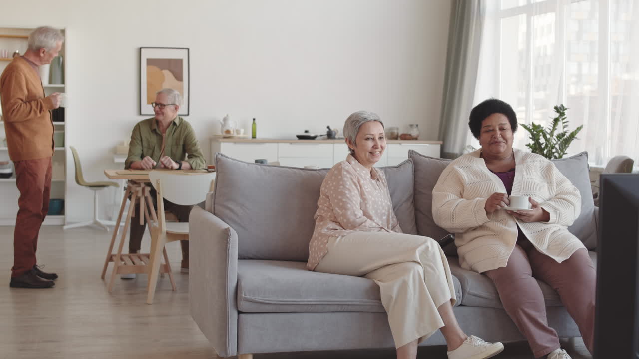 Breedbeeldopname van Aziatische en Afrikaanse oudere vrouwen die praten, op de bank zitten en tv kijken, met twee oudere mannen die op de achtergrond communiceren.