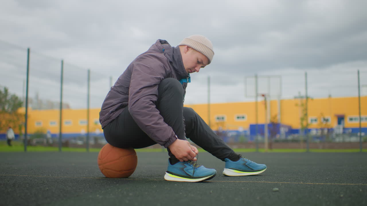 Açık hava sahasında spor ayakkabılarını bağlayan, basketbol beresi ve şişme mont üzerinde oturan bir atlet; soğuk, bulutlu bir gökyüzüne karşı, sarı bir bina ve çitli bir potanın arka planda yer aldığı ayakkabı bağlama işleminin yakın çekimi
