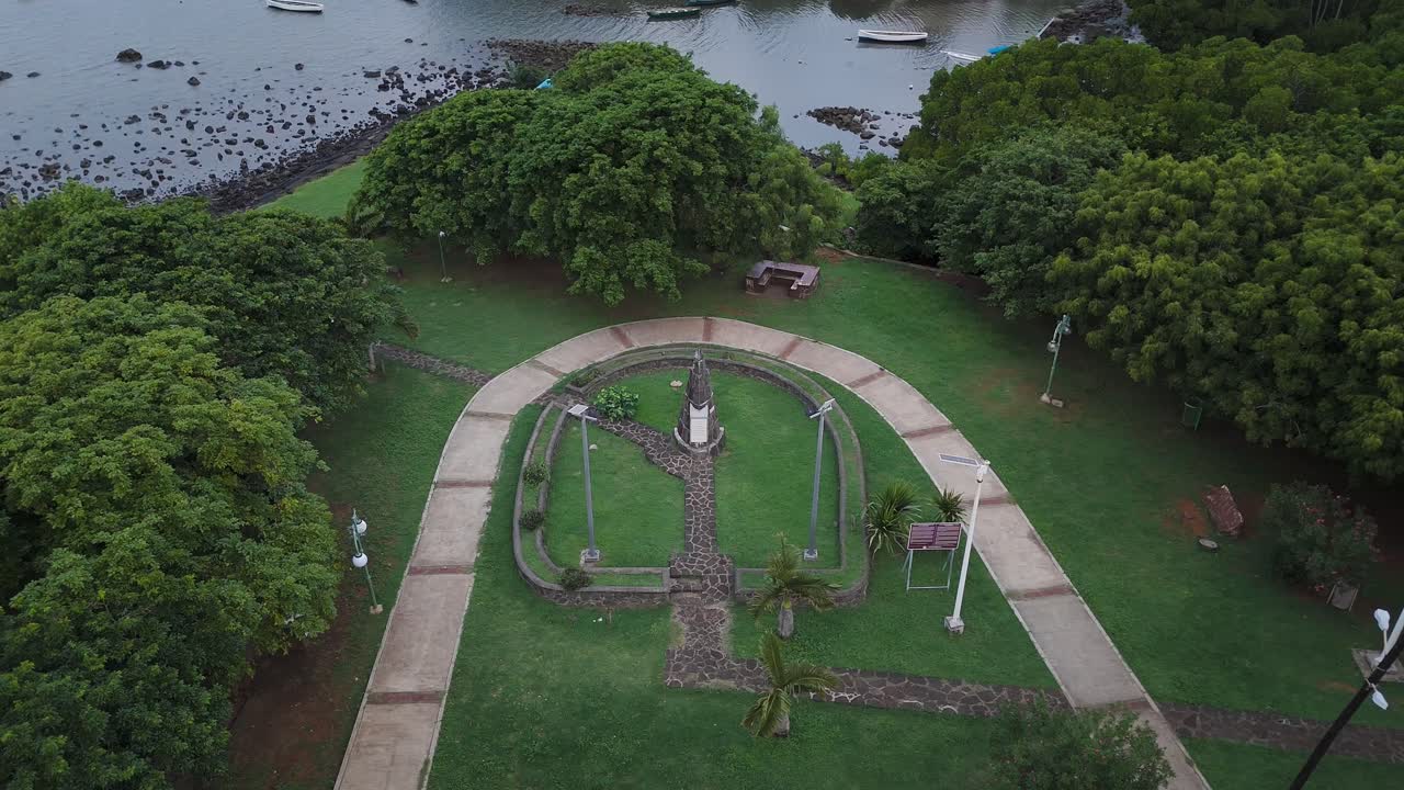 Aerial view of the Paul and Virginie Monument near Poudre d'Or Village, Mauritius, showing a coastal park, lush greenery, and boats along a serene shoreline under cloudy skies.
