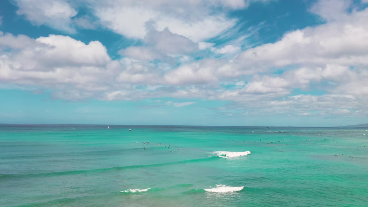 Surfers float on turquoise waves under bright skies off Waikīkī Beach, Oahu. Wide ocean view captures the serene beauty of Hawaii’s coastline with clouds drifting across the horizon