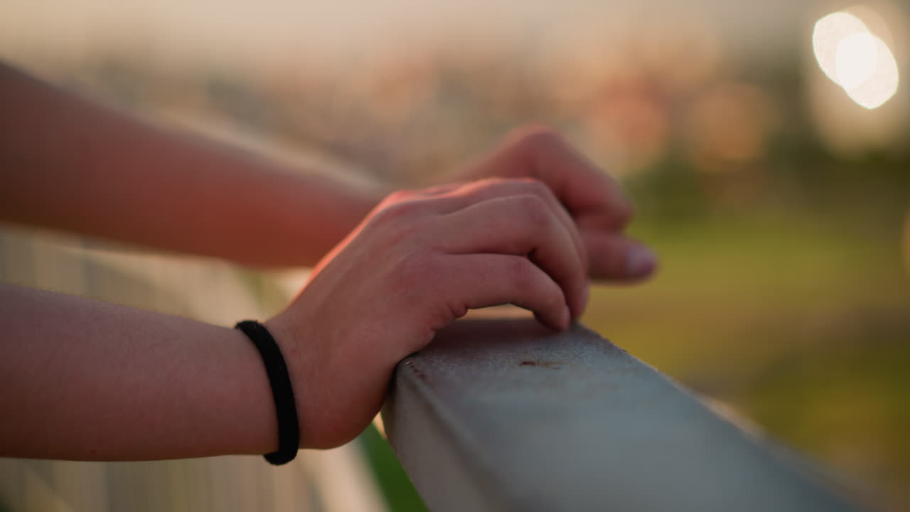 vista en primer plano de la mano de una persona blanca descansando en una barandilla de hierro con banda de muñeca negra en la mano derecha, fondo borroso con efecto de luz bokeh con colores cálidos