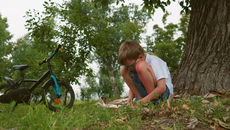 a thoughtful child squats under a tree with a red bruise on his leg, sitting on a blanket, a black bicycle with a blue front is parked nearby