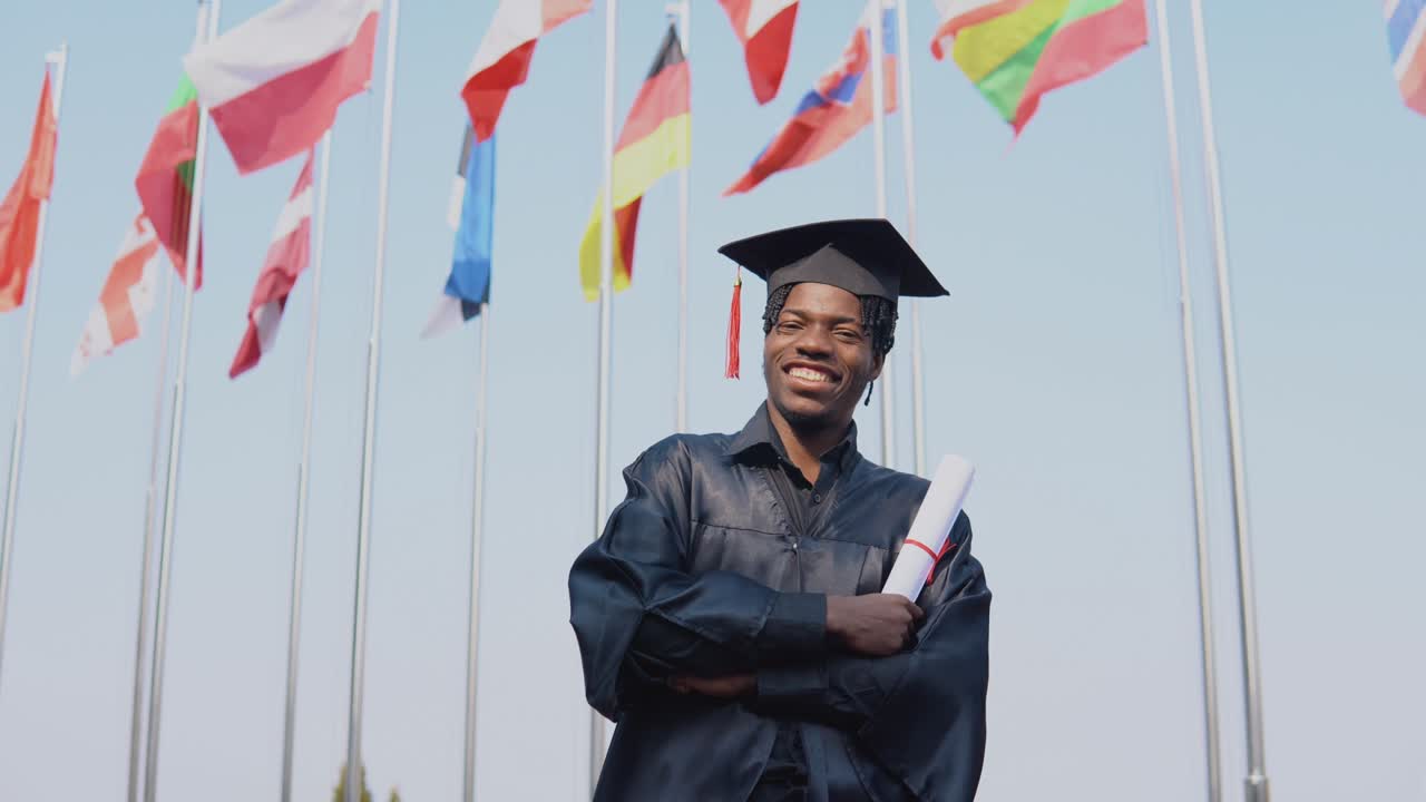 un joven graduado afroamericano feliz de pie frente a la cámara con un diploma en las manos. el estudiante está afuera con las banderas internacionales en el fondo.