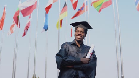 happy young african american male graduate standing in front of the camera with a diploma in his hands. the student stands outside with the international flags on background.