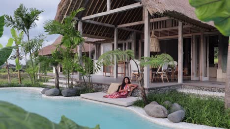 Cámara-Alejándose-De-Una-Joven-Modelo-Asiática-Sentada-Junto-A-La-Piscina-En-Una-Tumbona-Durante-El-Amanecer-De-La-Mañana-Con-Una-Lujosa-Villa-Boho-En-Medio-De-Un-Campo-De-Arroz-En-Bali,-Indonesia