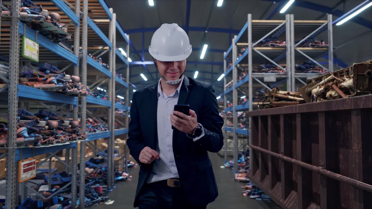 Vista frontale di un uomo d'affari asiatico che usa lo smartphone mentre cammina in un magazzino industriale di rottami metallici e riciclaggio.
