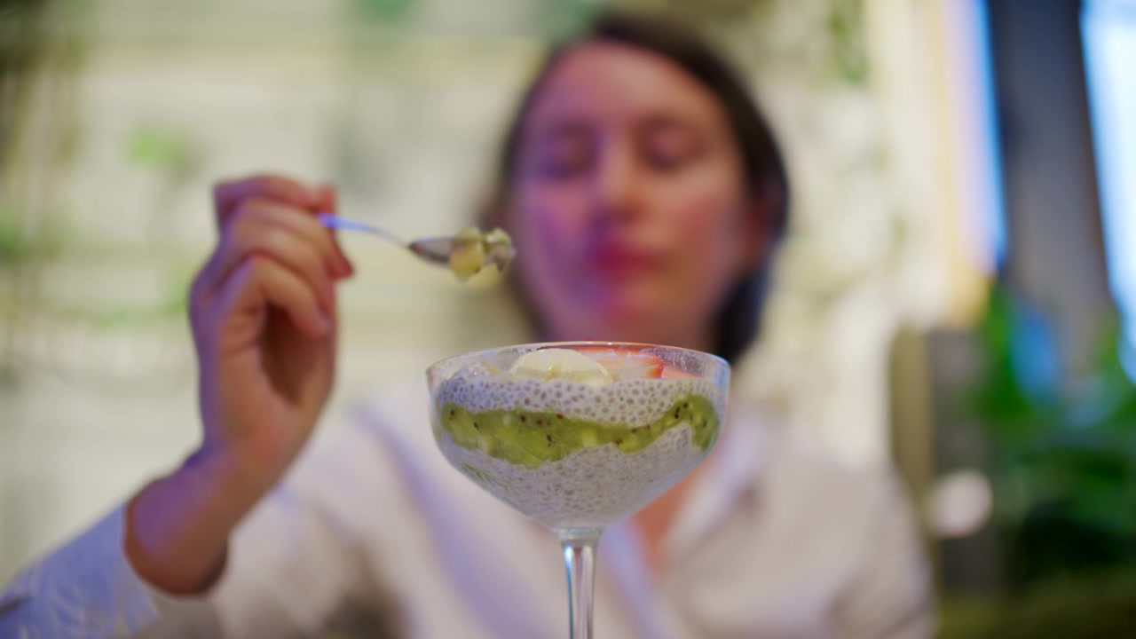 Bir kadın, beyaz chia tohumu pudingi, yeşil kivi püresi, dilimlenmiş çilek ve muzla dolu bir tatlı bardağının önünde bir kaşık chia pudingi tutuyor.