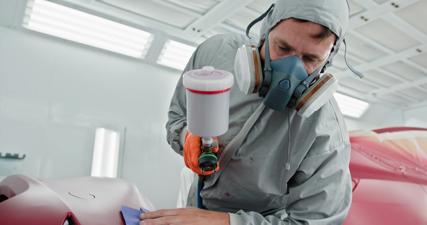 Técnico de carrocería con equipo de protección pintando con spray un parachoques de coche rojo en un taller de pintura especializado.