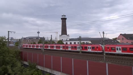 Cologne-Ehrenfeld,-Germany-2022---Red-commuter-train-passes-with-tower-in-the-background-on-a-cloudy-day