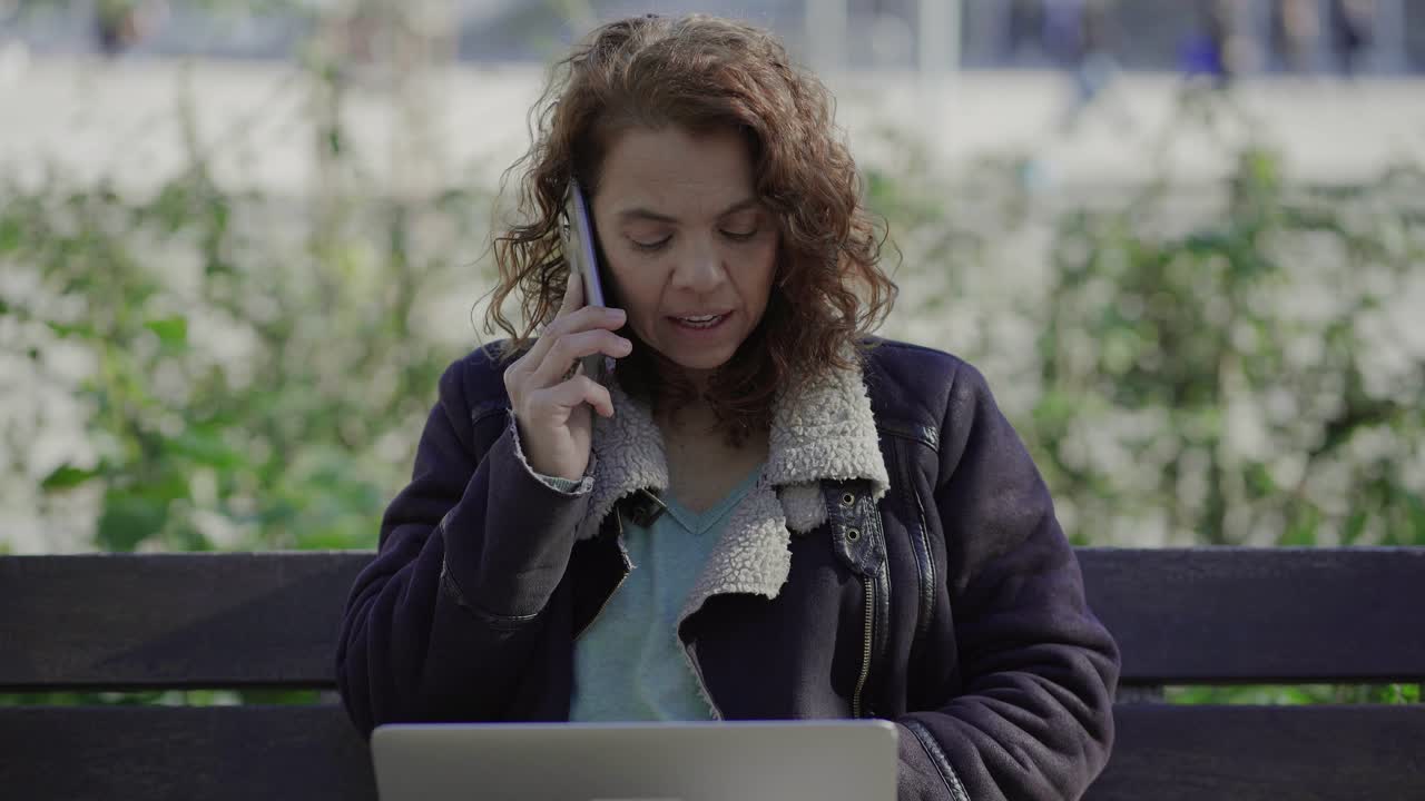 mujer rizada sonriente hablando en el teléfono inteligente mientras estaba sentada en el banco