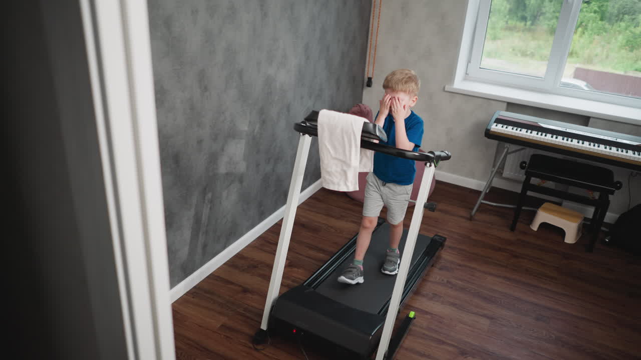 Cute boy walking on treadmill in cozy indoor space, scratching face and hand, focused expression, towel draped on handle, digital keyboard and stool in background, natural daylight through window