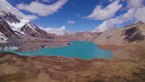 aerial view of tibetan plateau gorgeous blue colored lake between mountains that are brown and orange colored and have snow on the slopes during a sunny day. the sky is blue and has some clouds.