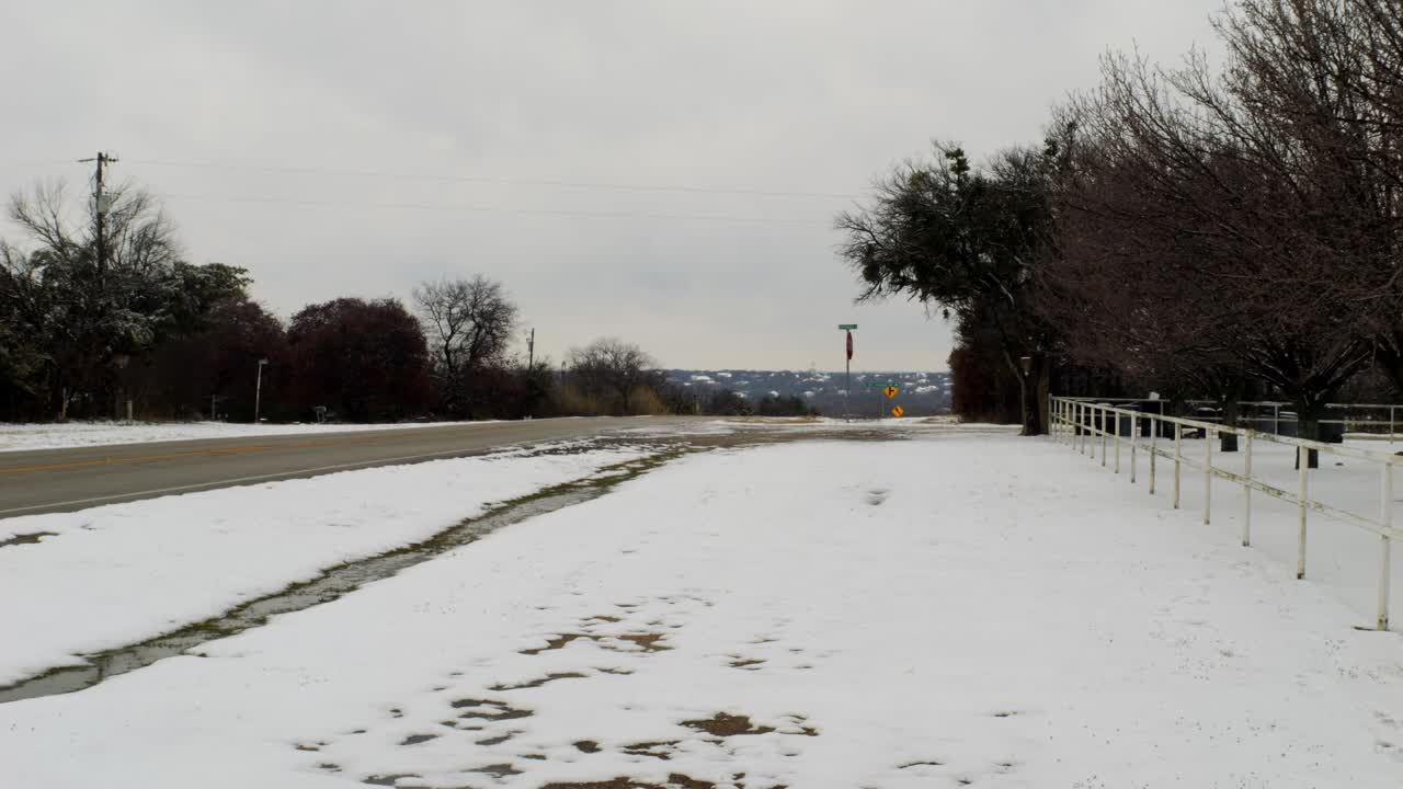 A car drives along a snowy roadside beside bare winter trees and distant hills, with light snow covering the ground and a calm, cold seasonal landscape