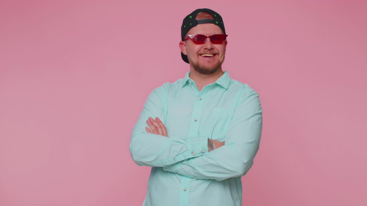 Verführerischer, fröhlicher, stilvoller Mann im blauen Hemd mit Sonnenbrille und charmantem Lächeln vor einer rosa Wand.