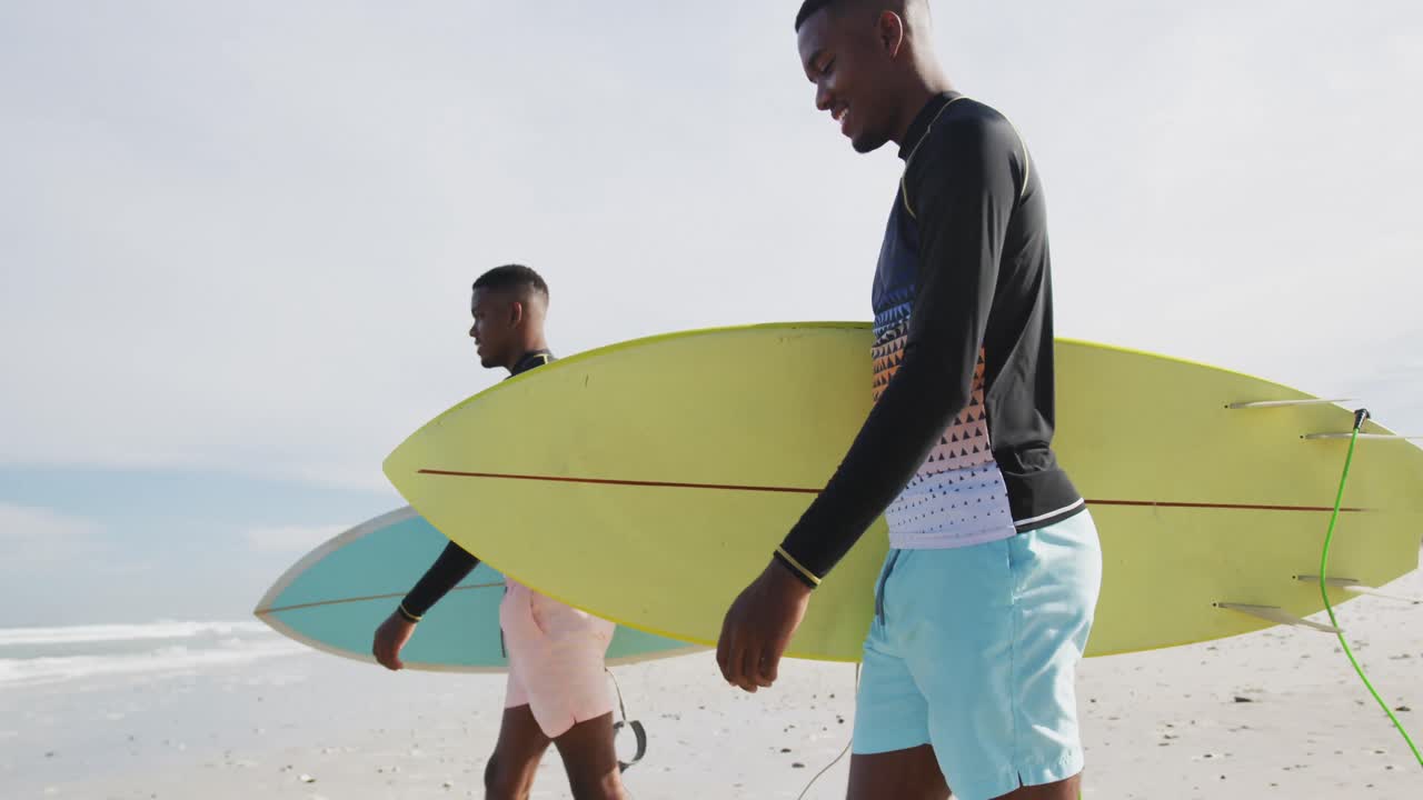 счастливые афроамериканские братья-близнецы-подростки гуляют по пляжу с досками для серфинга.