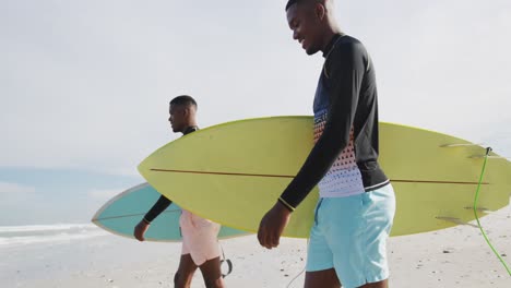 felices hermanos gemelos adolescentes afroamericanos caminando por una playa llevando tablas de surf