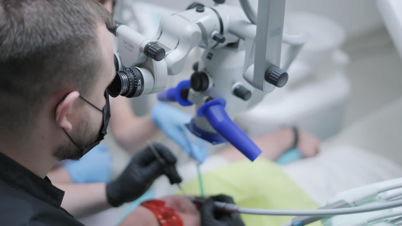 Male dentist working with dental microscope in modern stomatology clinic. Doctor using microscope for root canal treatment. Stomatologist performs a surgical operation. Cosmetic dentistry and surgery.