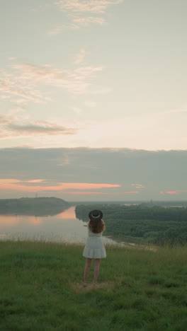 una mujer vestida con un vestido blanco y un sombrero se encuentra pensativa en un campo cubierto de hierba con vistas a un lago tranquilo al atardecer. mira hacia la distancia, creando una escena serena y reflexiva