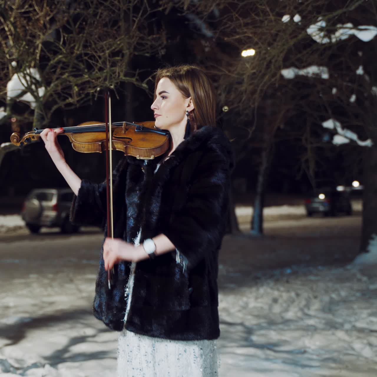 A beautiful girl with make-up and in a white dress is playing a violin composition in a winter park on the background of snow. Camera motion to left