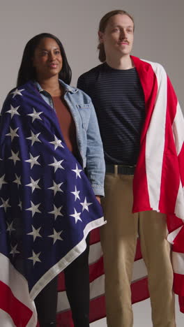retrato vertical de estudio de video de una pareja multicultural envuelta en la bandera estadounidense celebrando el 4 de julio, día de la independencia 1
