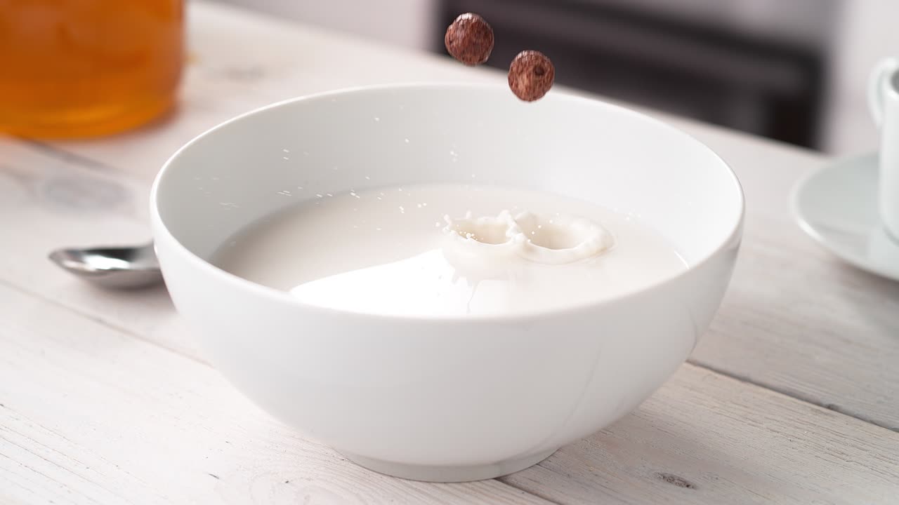 Cereais de chocolate em formato de bola caindo em tigela branca cheia de leite em câmera lenta.