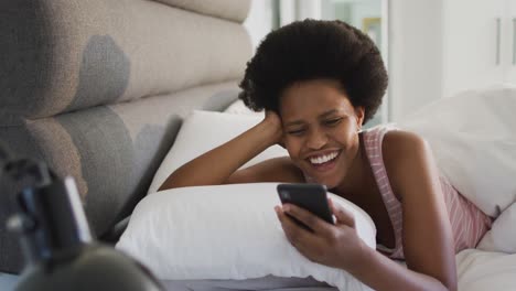 Happy-african-american-woman-laying-in-bed,-using-smartphone-in-bedroom