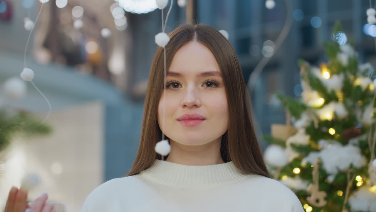 Young woman in white sweater gently interacts with a hanging white ornament in a mall, showcasing a playful and warm pose against a backdrop of festive decorations