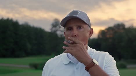 senior golf player smoking cigar looking camera on sunset field course fairway.