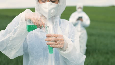 vista de cerca de una investigadora caucásica con traje protector que sostiene un tubo de ensayo mientras realiza el control de plagas en el campo verde