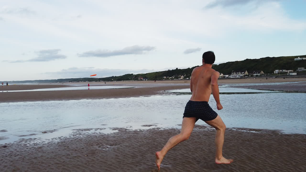 Shirtless man in shorts sprints barefoot across the wet sand of Omaha Beach to catch a frisbee, with tidal pools and coastal houses visible in the background, slow motion