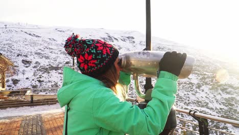 woman in a green jacket using binoculars at a viewpoint with a snowy landscape on a sunny day a reflection of the sun in the lens