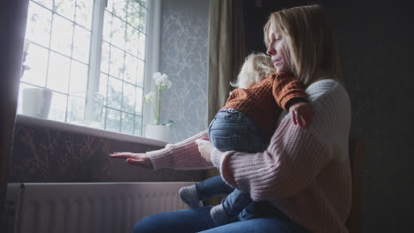 mother cuddling son trying to keep warm by radiator at home during cost of living energy crisis