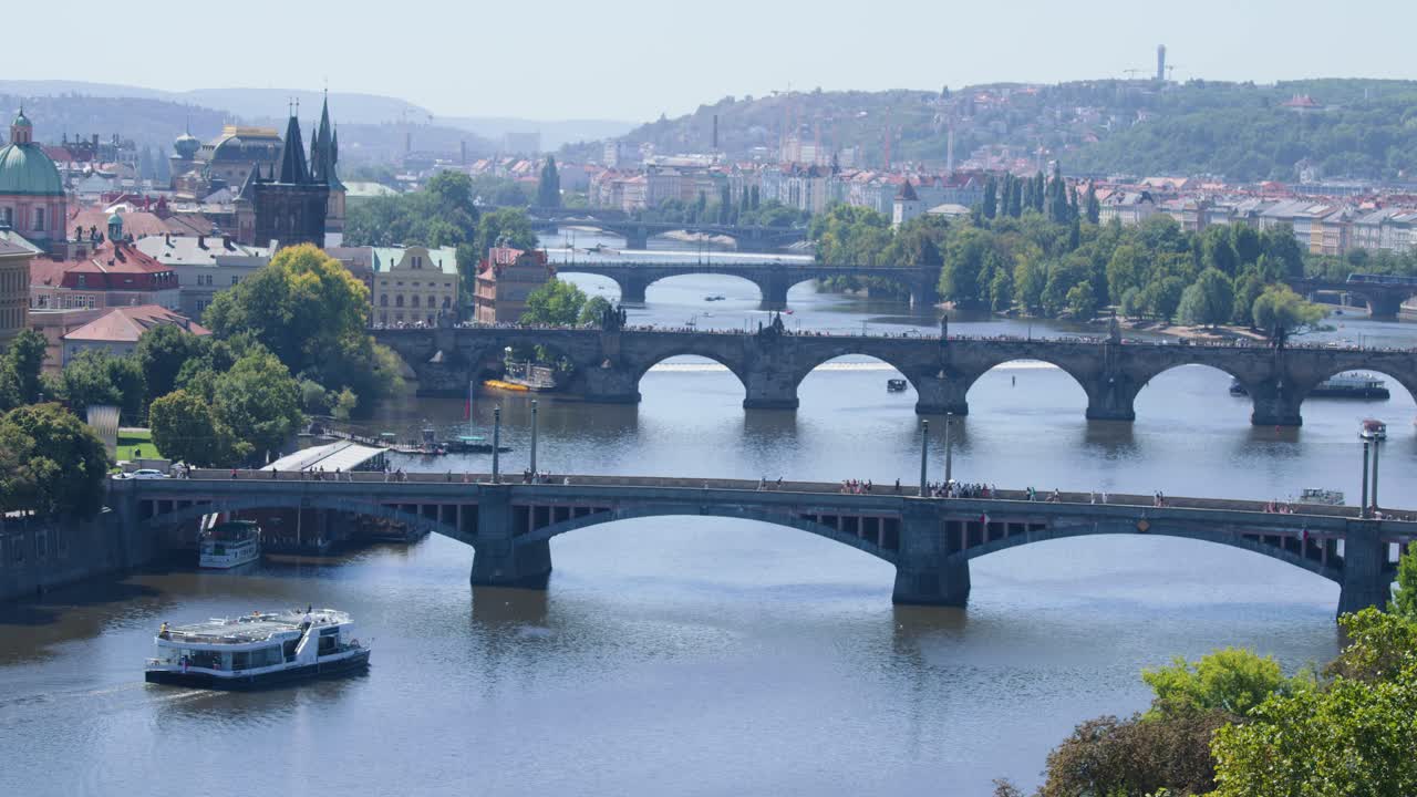 Sunny aerial shot of Prague’s Vltava River, historic bridges, cityscape, and moving tour boat
