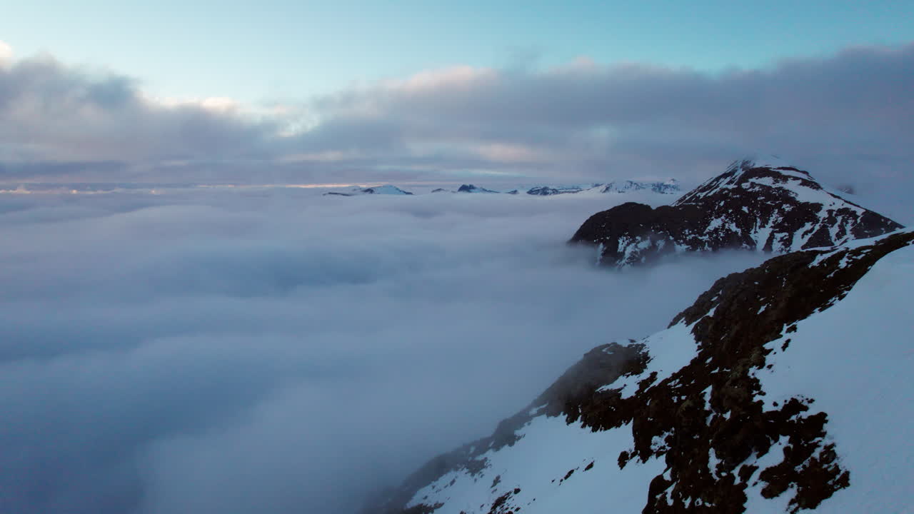 sobrevuelo aéreo de un montañero quitándose el suéter mientras mira por encima del borde de un valle cubierto de nubes y picos montañosos cubiertos de nieve se elevan con el sol bajo en el cielo